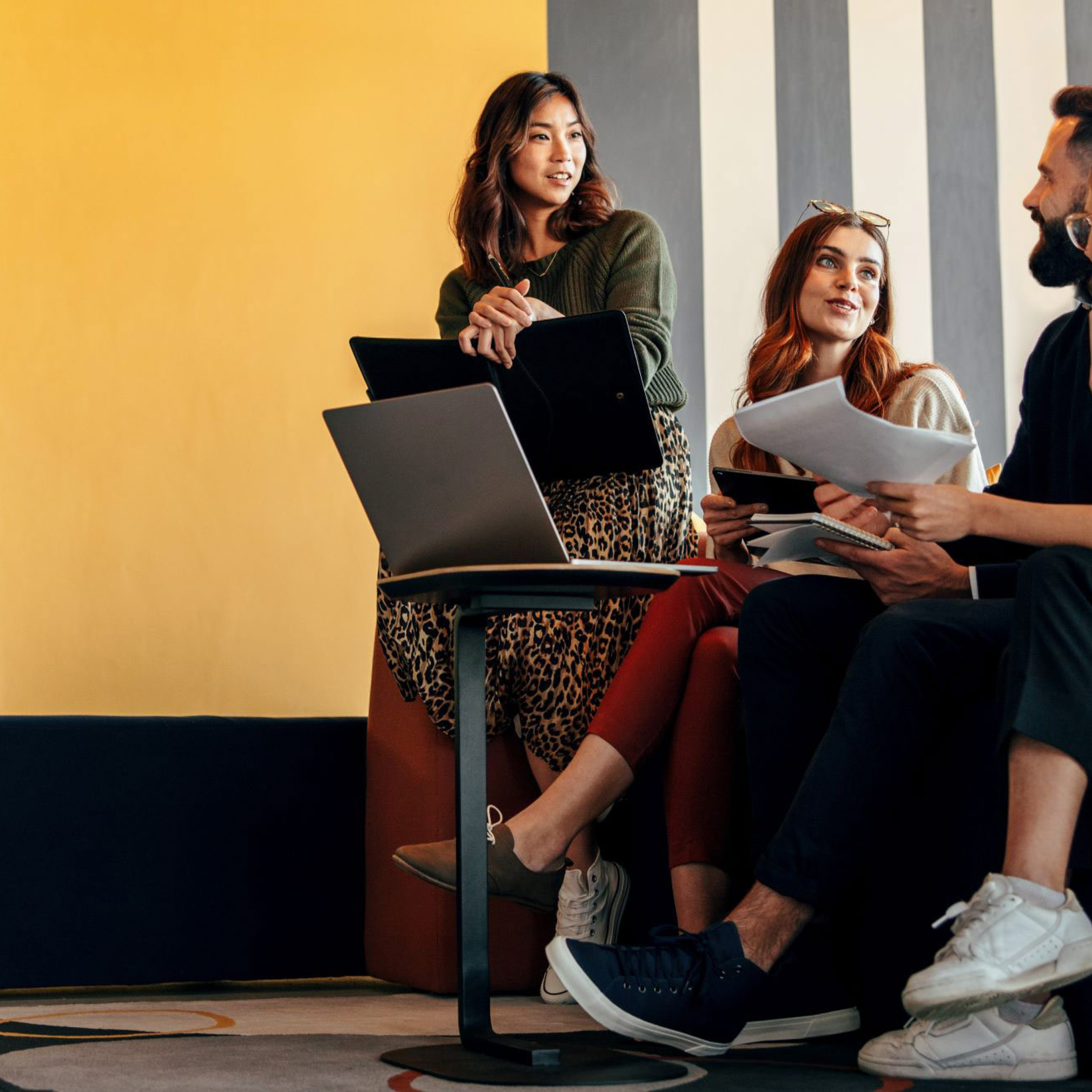 Four young adults sitting and talking in a modern workspace, with laptops and papers.
